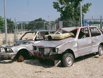 Zwei silberne Schrottautos, verbeult, teilweise ohne Reifen stehen vor einem Zaun auf einem Schotterpltz