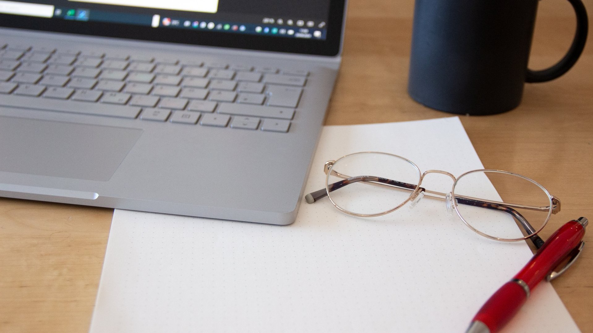 Brille, Stift, Block, Laptop und Kaffeetasse auf dem Schreibtisch.